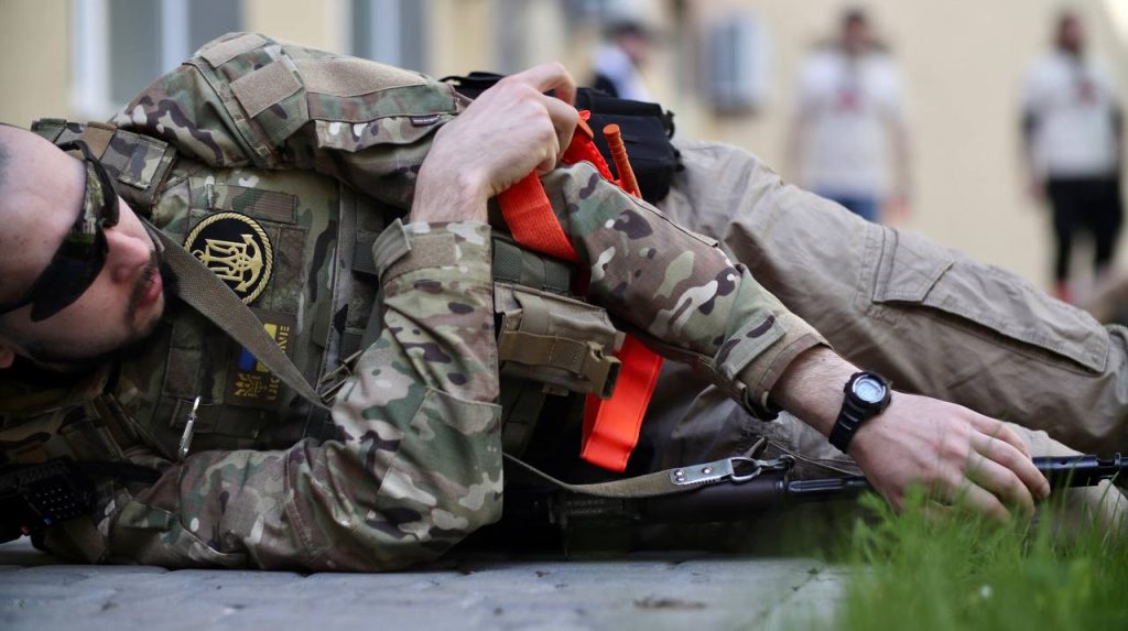 A man in military uniform is lying on the ground while practicing how to use a tourniquet