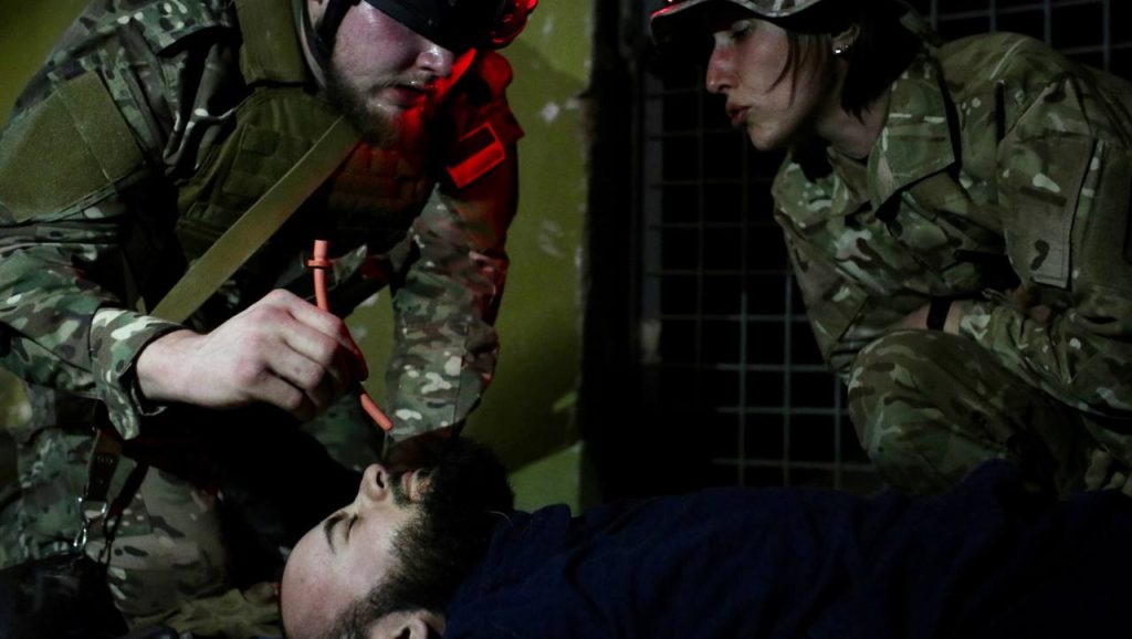 Two people in military uniforms are providing medical help to a man laying in front of them