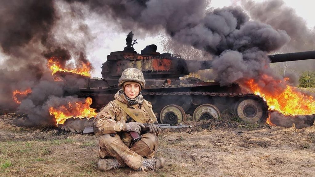 A woman in military uniform is sitting on the ground and holding a gun while a Russian tank is burning on the backdrop